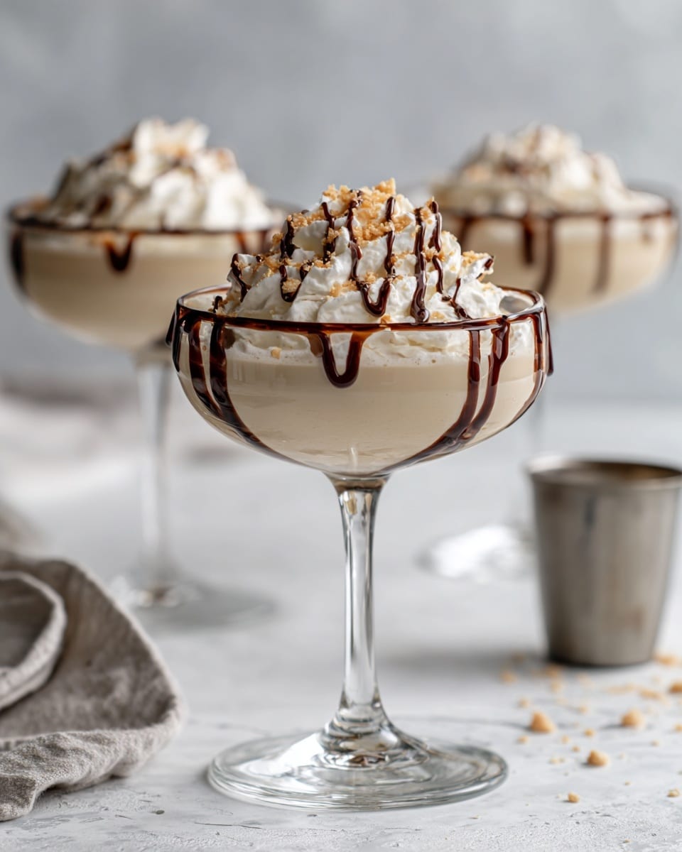 The image shows three white cocktail glasses each filled with a creamy light beige drink. Each glass is topped with a thick layer of white whipped cream, which is drizzled with dark brown chocolate sauce in thin, wavy lines. There are small pieces of light tan crushed nuts sprinkled on top, adding texture. The glasses have a wide bowl shape and a thin stem. The background is a white marbled texture with blurred shapes of bottles and a metal cup visible. Photo taken with an iphone --ar 4:5 --v 7