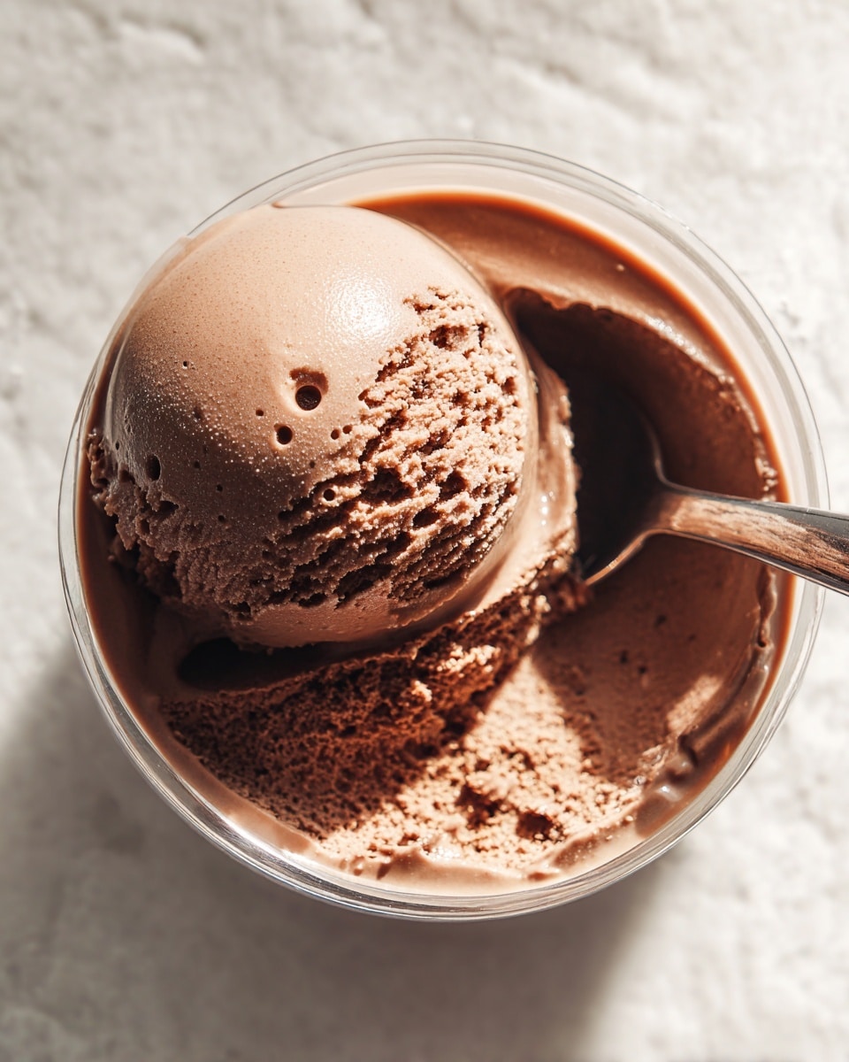 A close-up view of a single scoop of chocolate ice cream with a smooth and creamy texture, showing tiny air bubbles on its surface, sitting inside a clear glass or plastic round container full of the same chocolate ice cream. The ice cream has a rich brown color with a slight shine reflecting light on the top right side, and the container's inner edges have traces of melted chocolate ice cream. A metal spoon handle is placed inside the container on the right side, partially visible. The container rests on a white marbled surface. photo taken with an iphone --ar 4:5 --v 7