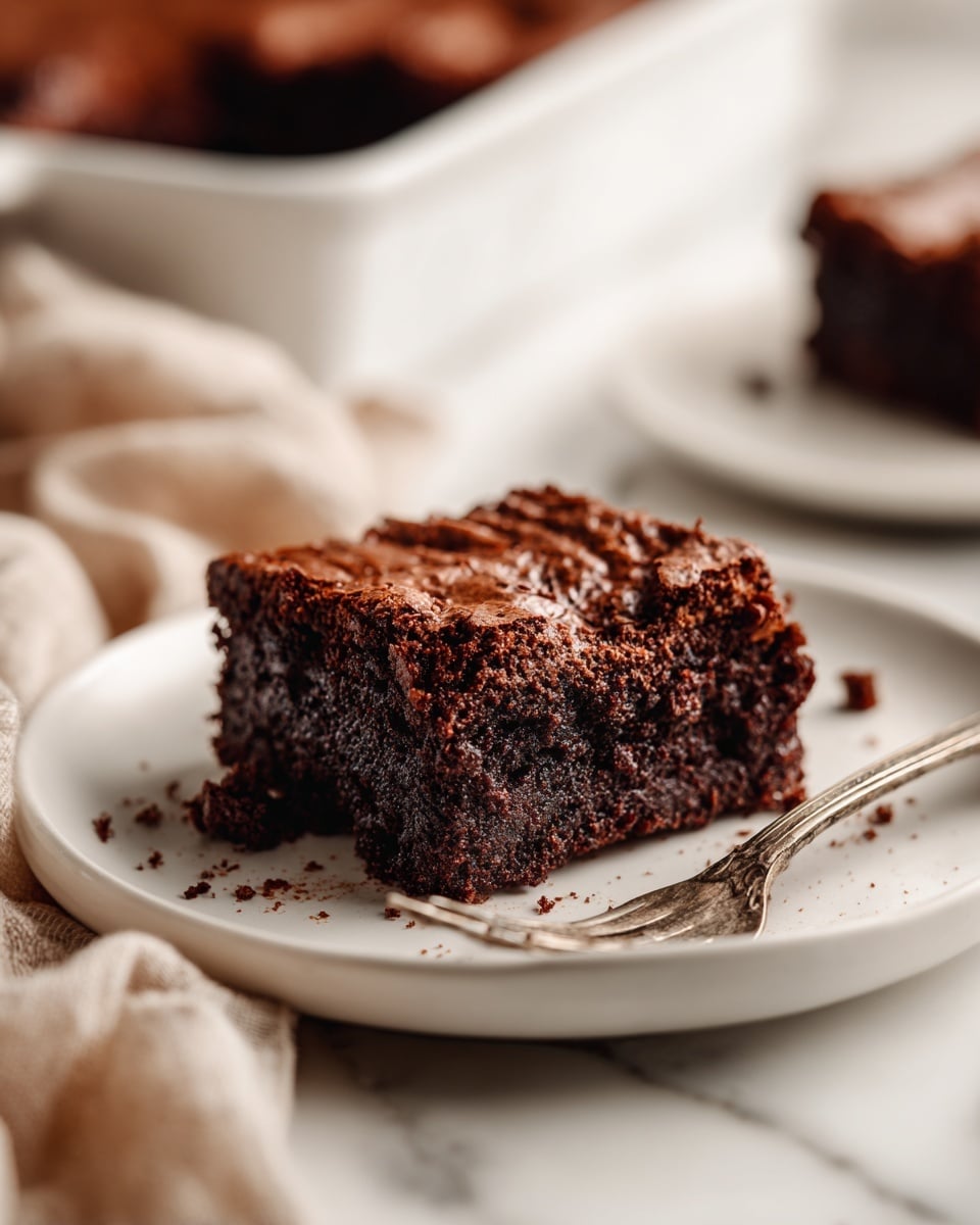 A single thick square piece of dark chocolate brownie with a moist and slightly crumbly texture sits on a white plate, showing a rich, dense interior with a shiny, slightly cracked top layer. The brownie has dark brown tones with uneven edges, and small crumbs are scattered around it on the plate. There is a vintage-style silver fork resting next to the brownie on the plate. In the blurred background, there is a white baking dish with more brownies, along with another piece of brownie on a white plate. All is set on a white marbled surface with a soft beige cloth nearby. Photo taken with an iphone --ar 4:5 --v 7