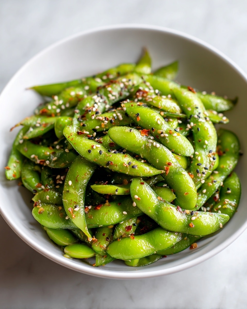 A close-up view of a white bowl filled with bright green edamame beans, some slightly open and others whole, all mixed with dark sesame seeds scattered evenly on top. The edamame beans have a fresh, smooth texture with a slight shine, and some red chili flakes add small spots of red color, creating a nice contrast. The bowl sits on a white marbled surface, and the photo captures the fresh, healthy look of the dish with vibrant green and a few dark and red highlights. photo taken with an iphone --ar 4:5 --v 7