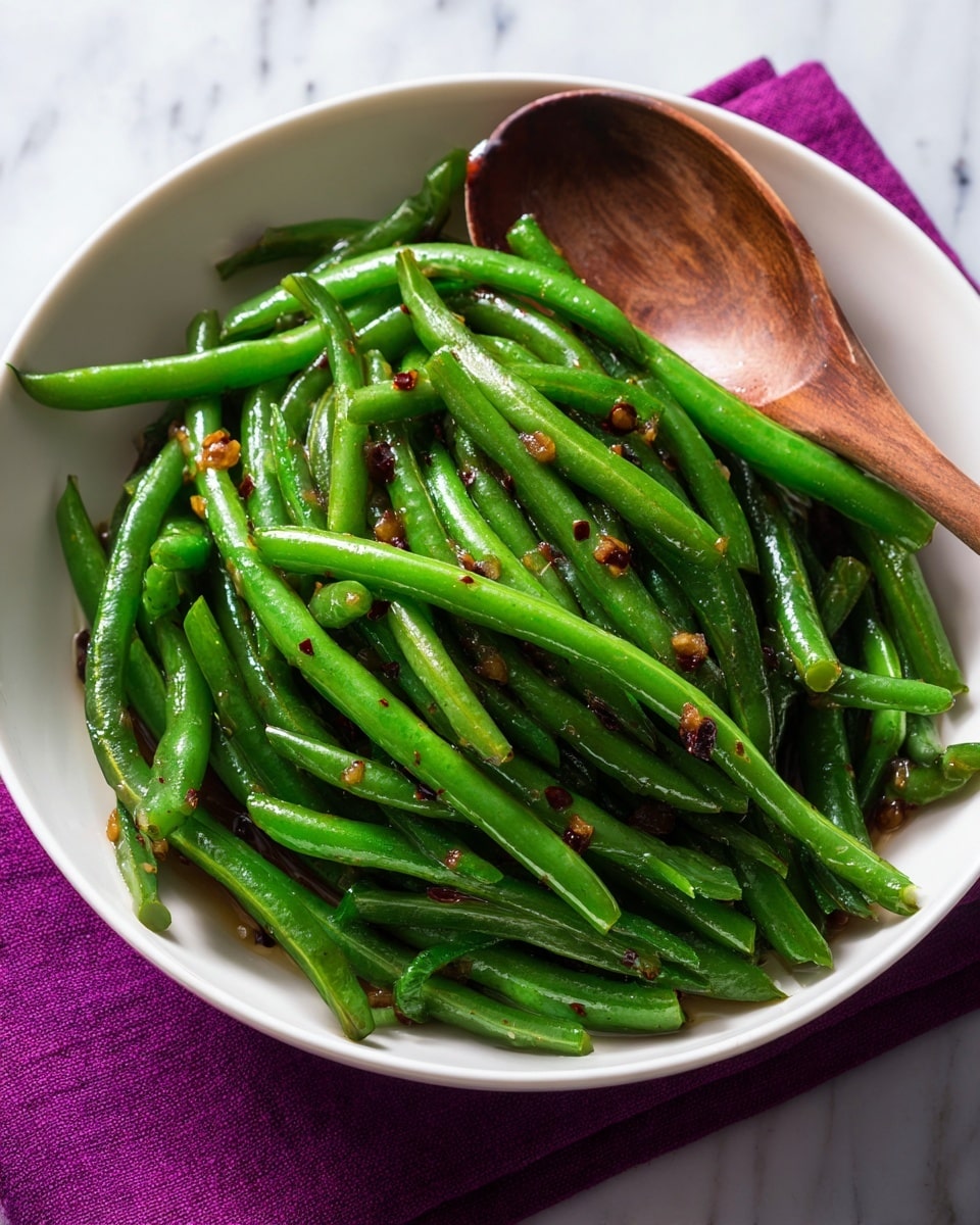 A bowl filled with a pile of shiny, cooked green beans mixed with small bits of browned garlic or onions, giving the green beans a slightly glossy look with some golden brown highlights. The green beans are long and placed in layers, with the top layer catching most of the light, showing their fresh green color and smooth texture. The bowl is white and sits on a white marbled surface, and a wooden spoon with a smooth finish rests inside the bowl on the right side, partially covered by the green beans. photo taken with an iphone --ar 4:5 --v 7