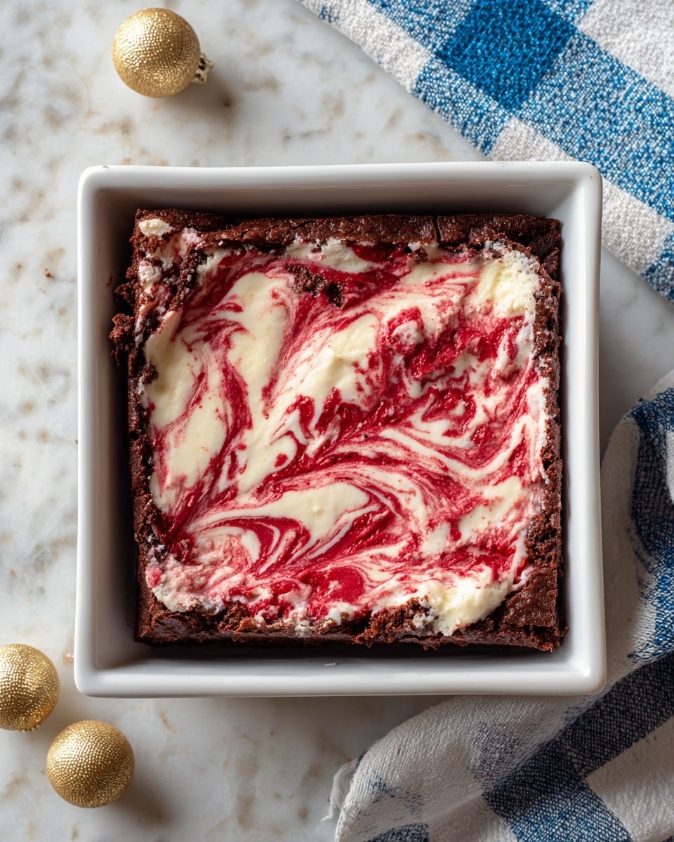 A square dessert sits in a white square baking dish on a white marbled surface. The dessert has two main layers: the bottom layer is a dark, rich chocolate brownie with a slightly cracked texture around the edges, and the top layer is a creamy swirl of red and white that looks like a mix of red velvet and cream cheese, creating a marbled pattern with smooth and rough textures. The edges of the brownie are thick and firm, contrasting with the soft, swirled top. Two small shiny gold balls are placed near the baking dish on the marbled surface, and two pieces of blue and white checkered cloth are partially visible on the top corners. Photo taken with an iphone --ar 4:5 --v 7