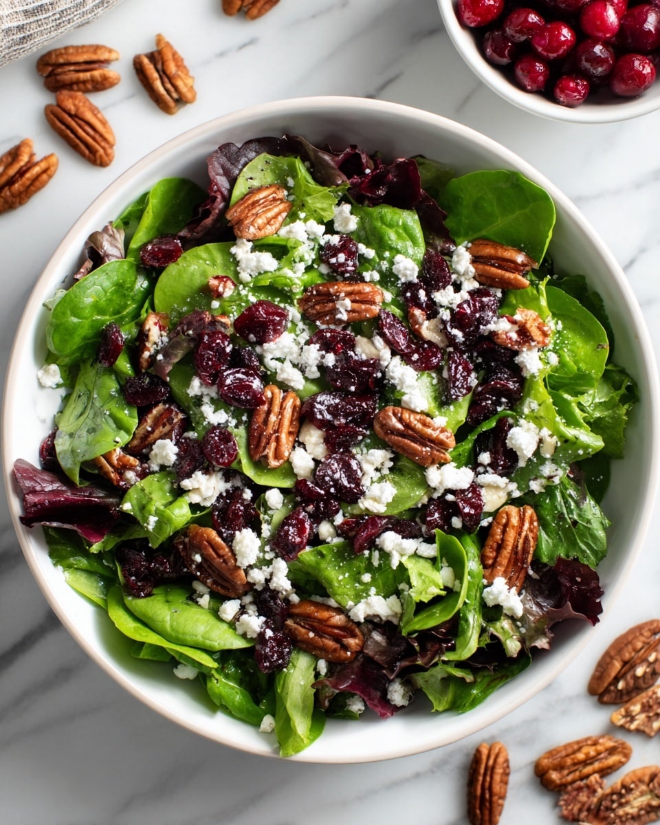 A fresh salad is shown in a white bowl on a white marbled surface, filled with bright green leafy mix including spinach and other greens as the base layer. On top of the leaves are small, deep red dried cranberries scattered evenly, followed by bits of crunchy light brown pecans. The salad is sprinkled with crumbled white cheese, adding a creamy texture contrast. In the background, there is a white bowl with more dried cranberries visible, and some whole pecans are placed casually on the surface near the bowl. photo taken with an iphone --ar 4:5 --v 7