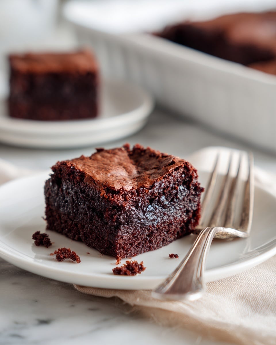 A square piece of dark chocolate brownie with a glossy cracked top layer sits on a white plate. The rich, moist texture is visible inside with small crumbs around the base. The plate rests on a light cream cloth over a white marbled surface. A silver fork lies next to the brownie on the plate. In the blurred background, another piece of brownie and a white baking dish holding more brownies are faintly seen. Photo taken with an iphone --ar 4:5 --v 7