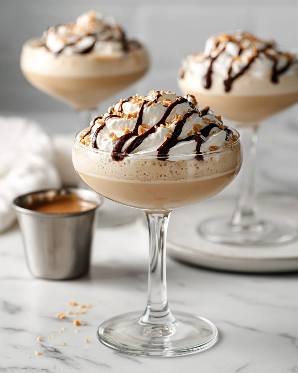 The image shows a creamy beige drink served in a white glass coupe, filled almost to the rim. On top, there is a thick layer of white whipped cream with a smooth, fluffy texture, decorated with a dark chocolate syrup drizzle in wavy lines and sprinkled with small light tan crumbs. In the background, two more identical glasses hold the same drink, slightly out of focus, creating depth. The setting is on a white marbled surface, with a small metal cup holding a similar liquid nearby. Photo taken with an iphone --ar 4:5 --v 7