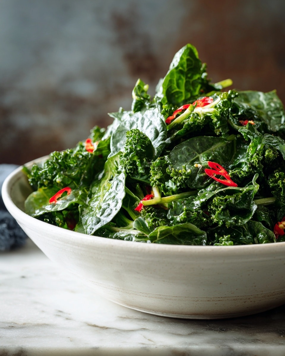 A large white bowl filled with dark green leafy kale and spinach leaves, layered thickly to create a fresh and hearty salad. The forward edges show larger spinach leaves with smooth texture while the deeper layers display curly and textured kale leaves. Bright red chili slices are scattered evenly throughout the greens, adding pops of vibrant color. Some light green stems poke out from within the leaves, giving a natural and fresh look. The bowl sits on a white marbled surface with a rustic dark blurred background, focusing attention on the fresh greens. Photo taken with an iphone --ar 4:5 --v 7