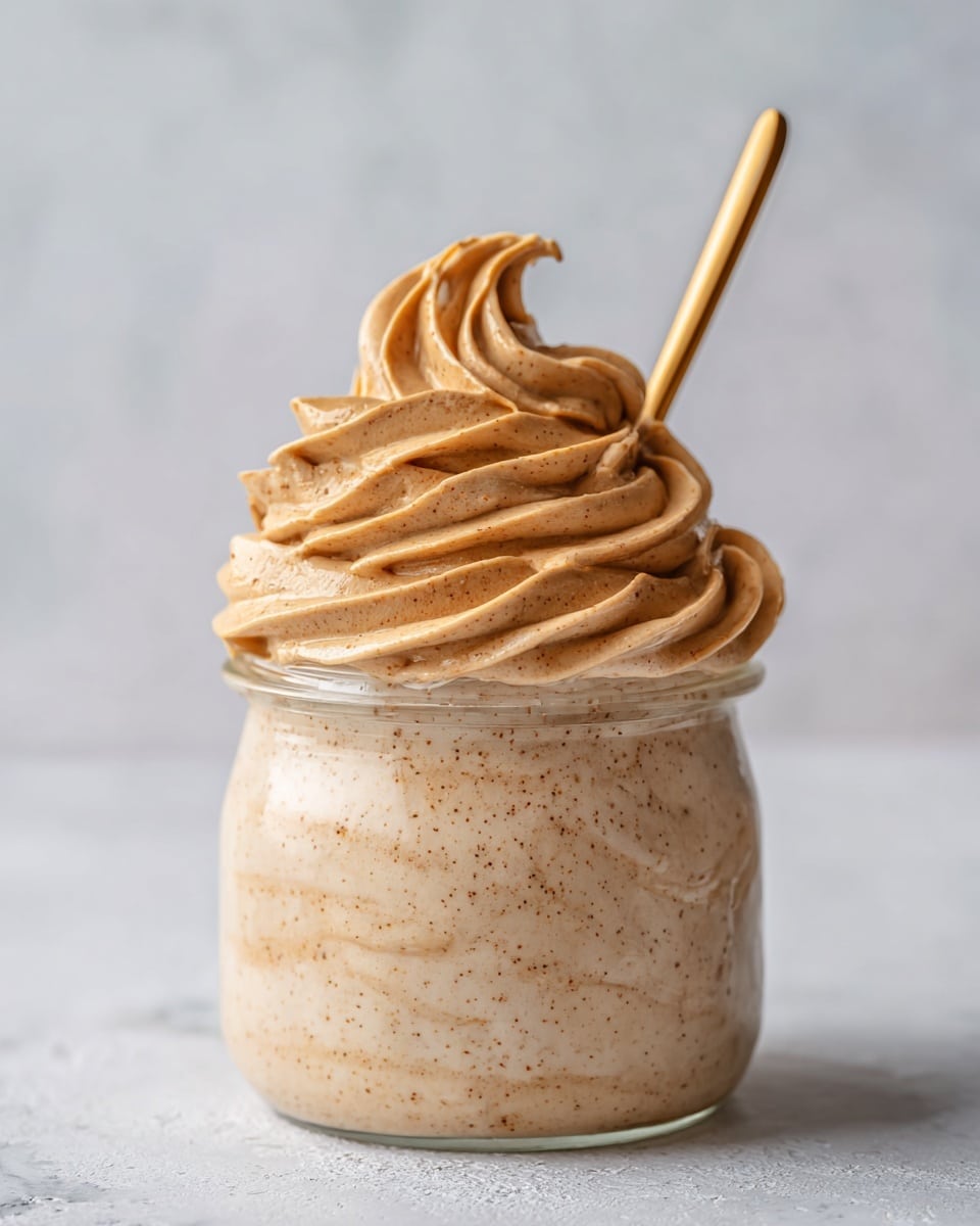 A clear glass jar filled with creamy peanut butter mousse that has a smooth, swirled texture on the top layer, light tan color, with some small peanut specks visible throughout. A gold spoon is partially dipped into the mousse, holding a small swirl in its bowl. The jar sits on a white marbled surface. photo taken with an iphone --ar 4:5 --v 7