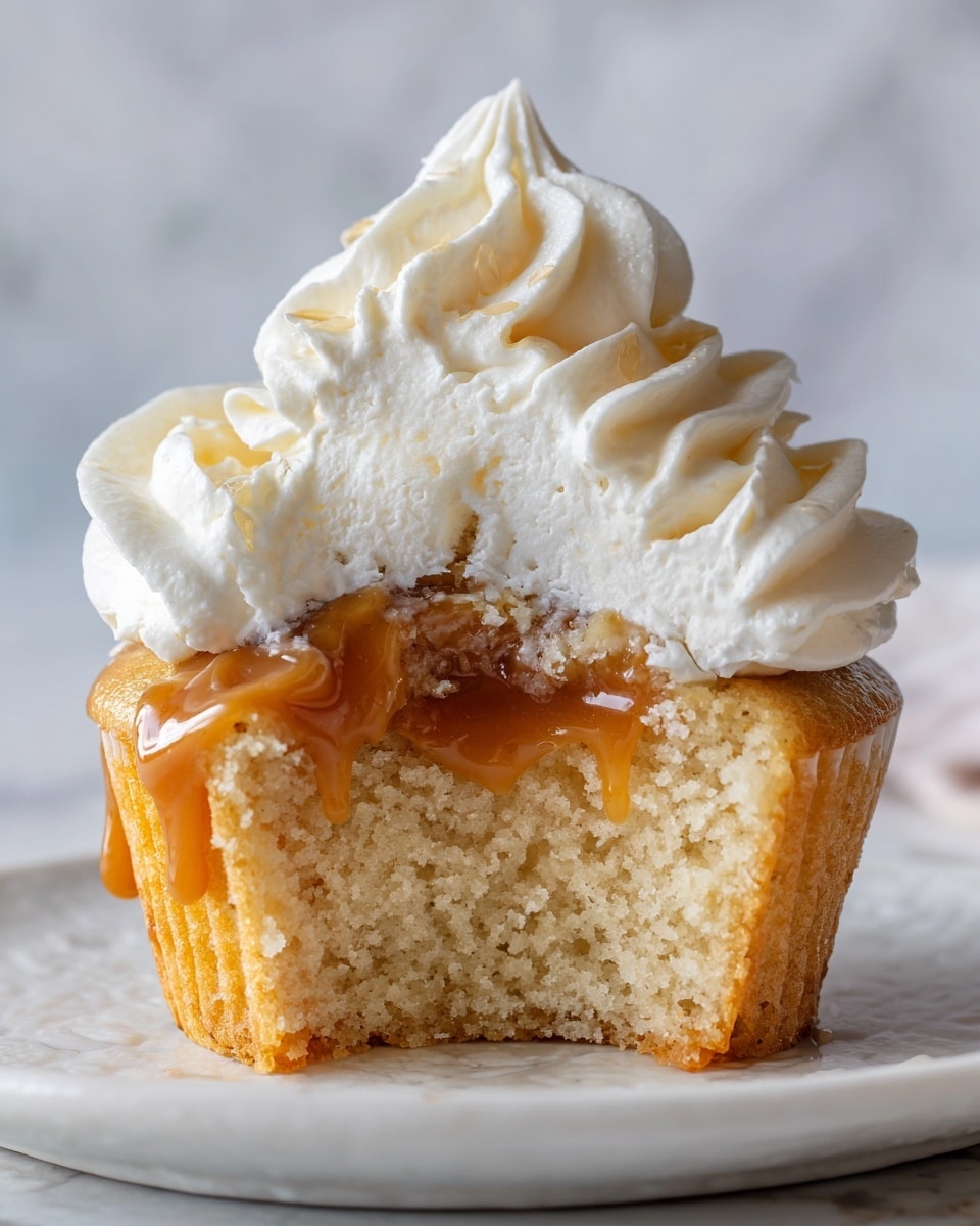 A close-up view of a vanilla cupcake with a bite taken out, showing three layers: a golden-brown, soft and crumbly cake forming the base, a creamy white frosting layer in the middle, and a glossy, smooth yellow caramel or custard center nestled within the frosting. The top of the cupcake is adorned with a fluffy swirl of white whipped cream, forming a delicate peak with soft, curvy edges. The cupcake sits on a white plate against a white marbled surface, with blurred cupcakes in the background. Photo taken with an iphone --ar 4:5 --v 7